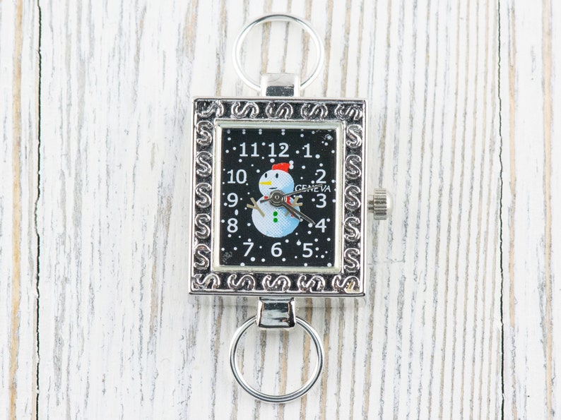 K&ouml;nnte beinhalten: Eine silberne Taschenuhr mit schwarzem Zifferblatt und einem Schneemann-Motiv. Die Uhr hat einen Schl&uuml;sselring.