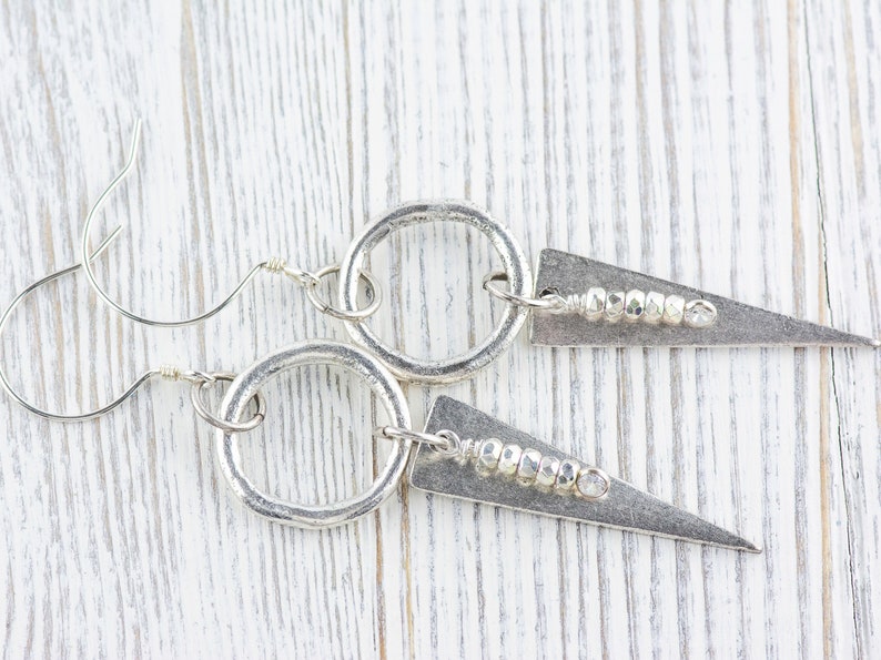 May include: Silver earrings with a circle and a triangle pendant. The triangle pendant has a row of small silver beads.