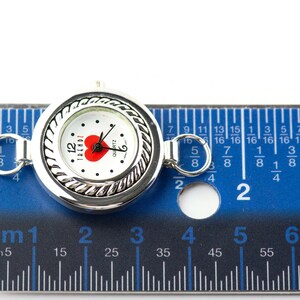 May include: A silver quartz watch with a white face, a red heart, and the word "TRENDZ". The watch is about 2.5 cm in diameter and has a decorative rope-like border. It is shown next to a blue ruler.