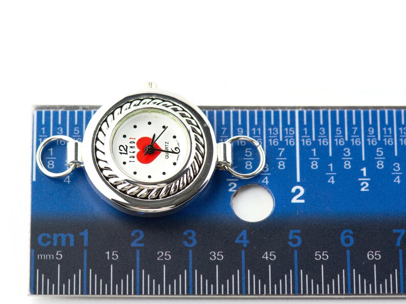 May include: A silver quartz watch with a white face, a red heart, and the word "TRENDZ". The watch is about 2.5 cm in diameter and has a decorative rope-like border. It is shown next to a blue ruler.