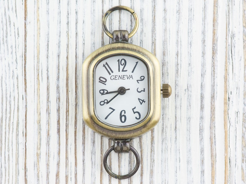 Pu&ograve; includere: Un orologio da tasca dorato con un quadrante quadrato e numeri romani. Il quadrante dell'orologio &egrave; bianco con numeri e lancette neri. L'orologio ha un piccolo anello in alto per attaccarlo a una catena.