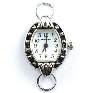 May include: Silver oval watch with a white face and black numerals. The watch has a decorative bezel with small, dark accents and a crown on the side. The watch face reads "NAEMI". Two silver rings are attached to the top and bottom.