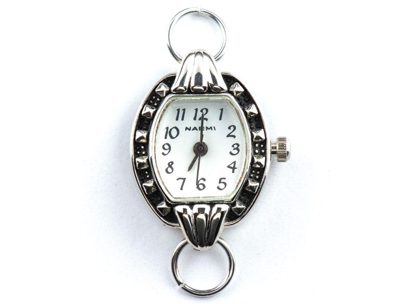 May include: Silver oval watch with a white face and black numerals. The watch has a decorative bezel with small, dark accents and a crown on the side. The watch face reads "NAEMI". Two silver rings are attached to the top and bottom.