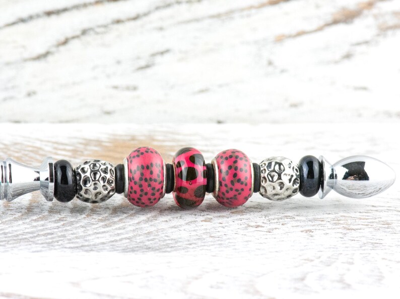 May include: A decorative bottle stopper with a silver finish and a series of beads. The beads include black, silver, and pink with black polka dots. The stopper is designed to fit into a bottle.