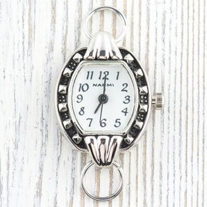 May include: Silver watch with a white face and black numerals. The watch has an oval frame with studded detailing and two ring attachments. The watch face reads "NAEMI".