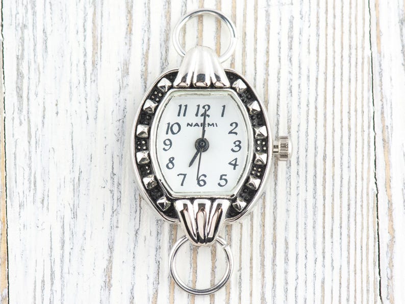 May include: Silver watch with a white face and black numerals. The watch has an oval frame with studded detailing and two ring attachments. The watch face reads "NAEMI".