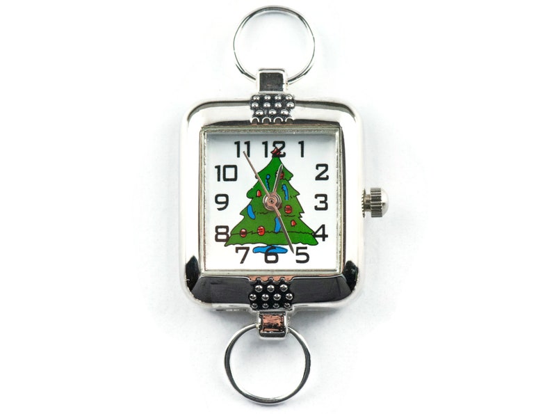 Puede incluir: Un reloj de llavero rectangular plateado con una esfera blanca con n&uacute;meros negros y un dise&ntilde;o de &aacute;rbol de Navidad verde. El reloj tiene una corona para ajustar la hora y dos llaveros.