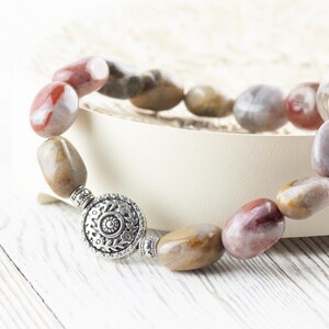Op de afbeelding: Een armband gemaakt van natuurlijke stenen kralen in tinten bruin, rood en wit. De kralen zijn ovaalvormig en hebben een gepolijste afwerking. De armband heeft een zilveren sluiting met een decoratief ontwerp.