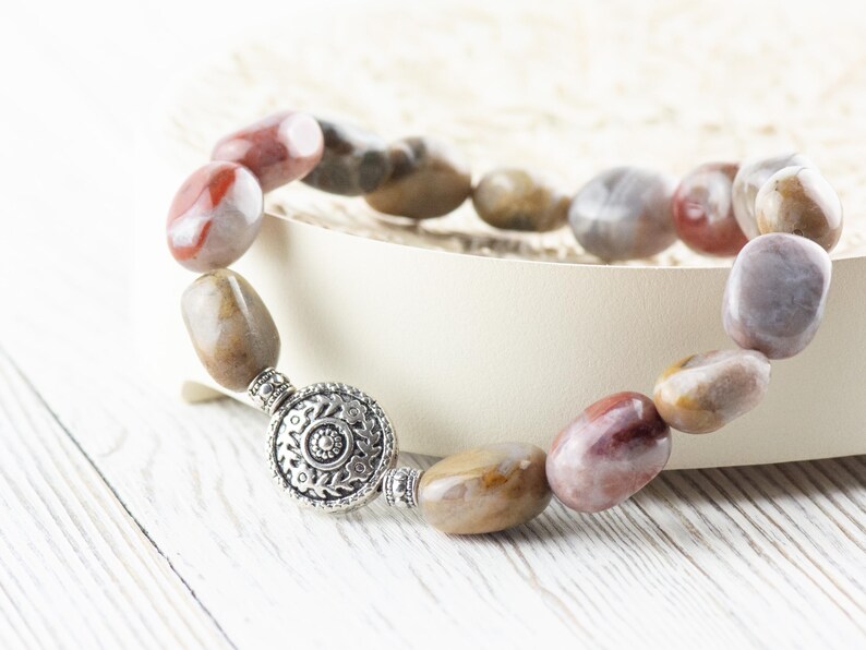 Op de afbeelding: Een armband gemaakt van natuurlijke stenen kralen in tinten bruin, rood en wit. De kralen zijn ovaalvormig en hebben een gepolijste afwerking. De armband heeft een zilveren sluiting met een decoratief ontwerp.