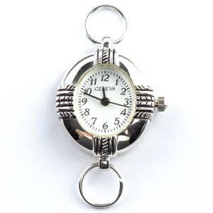 Pu&ograve; includere: Orologio portachiavi Geneva argentato con quadrante bianco e lancette nere. L'orologio ha un quadrante rotondo con numeri arabi e una lunetta strutturata. Include anelli nella parte superiore e inferiore per il fissaggio.