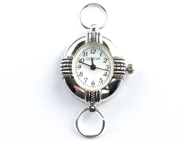 Pu&ograve; includere: Orologio portachiavi Geneva argentato con quadrante bianco e lancette nere. L'orologio ha un quadrante rotondo con numeri arabi e una lunetta strutturata. Include anelli nella parte superiore e inferiore per il fissaggio.