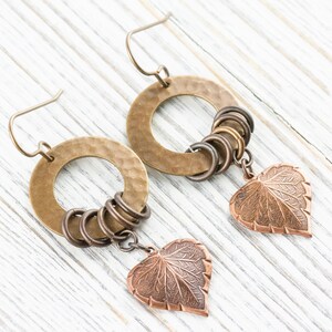 Op de afbeelding: Een paar handgemaakte oorbellen met gehamerde messing hoepels, kleinere bronzen ringen en koperen bladbedels. De oorbellen hebben een rustieke, bohemien stijl en worden gepresenteerd op een wit houten oppervlak.