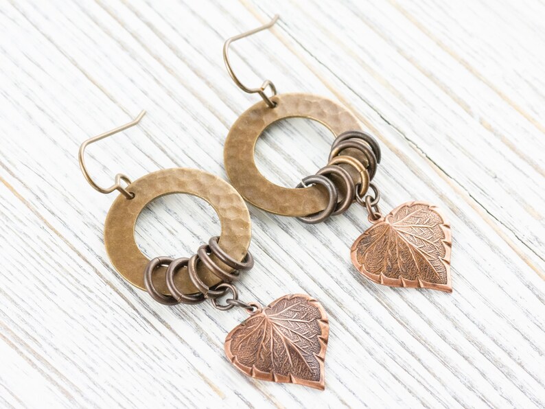 Op de afbeelding: Een paar handgemaakte oorbellen met gehamerde messing hoepels, kleinere bronzen ringen en koperen bladbedels. De oorbellen hebben een rustieke, bohemien stijl en worden gepresenteerd op een wit houten oppervlak.