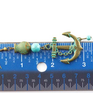 Puede incluir: Un par de pendientes colgantes con un dije de ancla azul turquesa y bronce, una cuenta azul turquesa y un dije de pez de bronce. Los pendientes se miden con una regla.