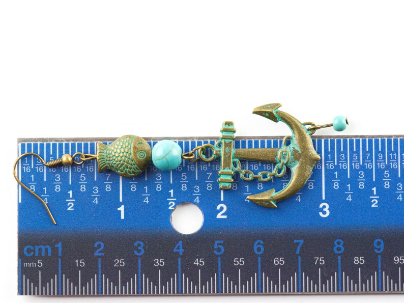 Puede incluir: Un par de pendientes colgantes con un dije de ancla azul turquesa y bronce, una cuenta azul turquesa y un dije de pez de bronce. Los pendientes se miden con una regla.