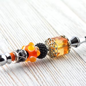 May include: A decorative letter opener with a silver blade and a handle adorned with colorful beads. The handle features orange, black, and silver beads, including a large orange and yellow bead and a filigree-wrapped amber-colored bead.