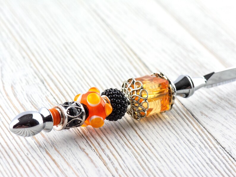 May include: A decorative letter opener with a silver blade and a handle adorned with colorful beads. The handle features orange, black, and silver beads, including a large orange and yellow bead and a filigree-wrapped amber-colored bead.