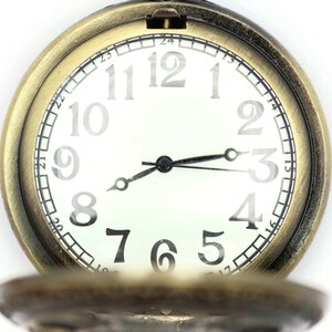 K&ouml;nnte beinhalten: Eine goldfarbene Taschenuhr mit wei&szlig;em Zifferblatt und schwarzen Zeigern. Das Zifferblatt der Uhr hat schwarze r&ouml;mische Ziffern f&uuml;r die Stunden und schwarze Markierungen f&uuml;r die Minuten.