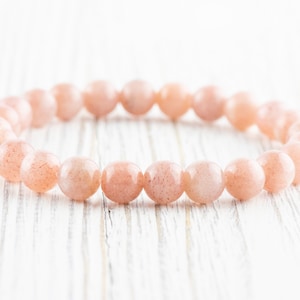 May include: A bracelet made of light pink sunstone beads. The beads are round and smooth, and the bracelet is strung on a clear elastic cord.