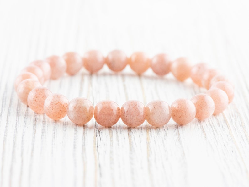 May include: A bracelet made of light pink sunstone beads. The beads are round and smooth, and the bracelet is strung on a clear elastic cord.