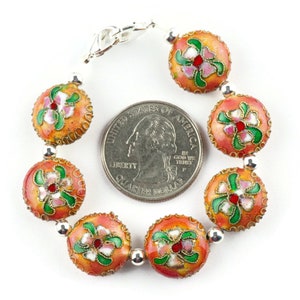 May include: A bracelet made of round, orange beads with floral designs in pink, green, and white. The beads are about the size of a US quarter. The bracelet has a silver clasp.