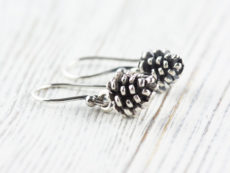 May include: A pair of silver dangle earrings with a pine cone design. The pine cones are detailed and realistic.