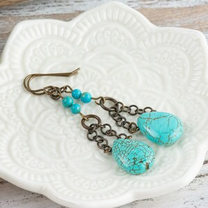 May include: Turquoise dangle earrings with a chain and beads. The earrings are on a white ceramic dish with a floral design.