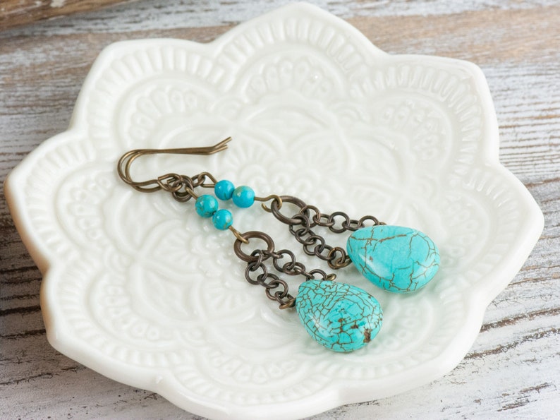 May include: Turquoise dangle earrings with a chain and beads. The earrings are on a white ceramic dish with a floral design.