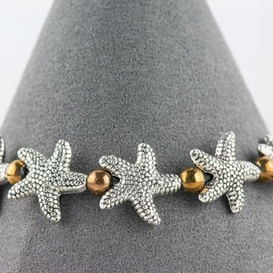 K&ouml;nnte beinhalten: Ein silbernes Armband mit einem sich wiederholenden Muster aus Seesternen und goldenen Perlen.