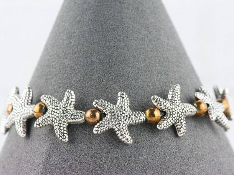 K&ouml;nnte beinhalten: Ein silbernes Armband mit einem sich wiederholenden Muster aus Seesternen und goldenen Perlen.
