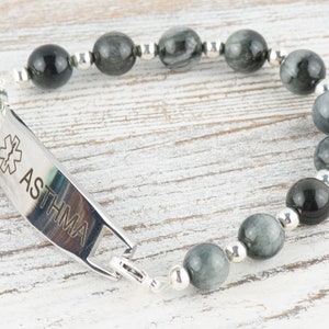 May include: A silver medical alert bracelet with a black and grey tiger's eye bead bracelet. The medical alert tag is engraved with a medical symbol and the word "ASTHMA".