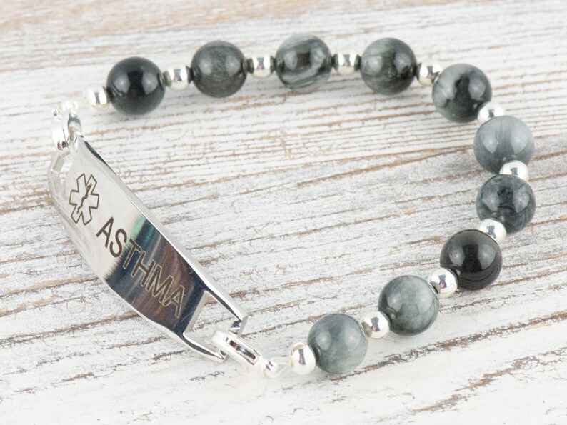 May include: A silver medical alert bracelet with a black and grey tiger's eye bead bracelet. The medical alert tag is engraved with a medical symbol and the word "ASTHMA".