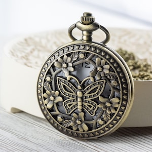K&ouml;nnte beinhalten: Antike bronzefarbene Taschenuhr mit einem Schmetterlingsdesign auf dem Zifferblatt, umgeben von floralen Akzenten. Die Uhr hat eine Kette und eine detaillierte, dekorative L&uuml;nette. Die Uhr ist vor einem neutralen Hintergrund platziert.