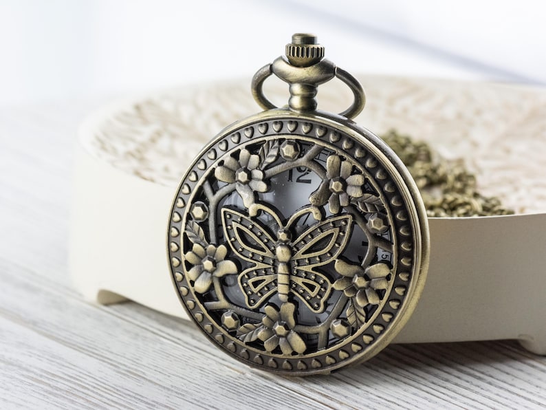 K&ouml;nnte beinhalten: Antike bronzefarbene Taschenuhr mit einem Schmetterlingsdesign auf dem Zifferblatt, umgeben von floralen Akzenten. Die Uhr hat eine Kette und eine detaillierte, dekorative L&uuml;nette. Die Uhr ist vor einem neutralen Hintergrund platziert.