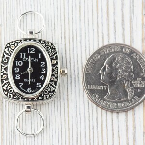 K&ouml;nnte beinhalten: Eine silberne Taschenuhr mit schwarzem Zifferblatt und quadratischem Geh&auml;use. Die Uhr ist mit einem verzierten Design verziert und hat einen kleinen Ring an der Seite. Die Uhr befindet sich neben einem US-amerikanischen Viertel f&uuml;r den Gr&ouml;&szlig;envergleich.