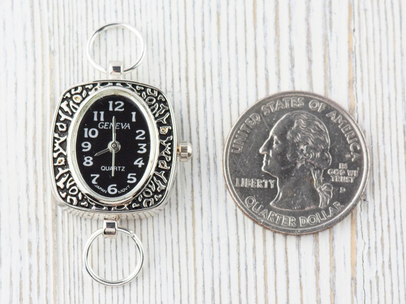 K&ouml;nnte beinhalten: Eine silberne Taschenuhr mit schwarzem Zifferblatt und quadratischem Geh&auml;use. Die Uhr ist mit einem verzierten Design verziert und hat einen kleinen Ring an der Seite. Die Uhr befindet sich neben einem US-amerikanischen Viertel f&uuml;r den Gr&ouml;&szlig;envergleich.