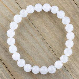 May include: A white beaded bracelet made of round, smooth stones. The bracelet is on a wooden surface.
