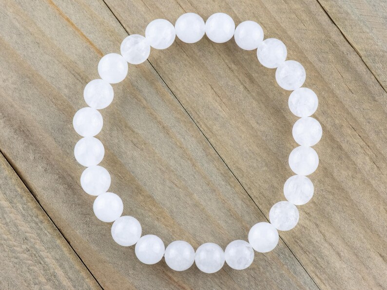 May include: A white beaded bracelet made of round, smooth stones. The bracelet is on a wooden surface.