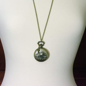 Pode incluir: Um colar dourado com um pingente de rel&oacute;gio de bolso redondo que apresenta um design de peixe detalhado.