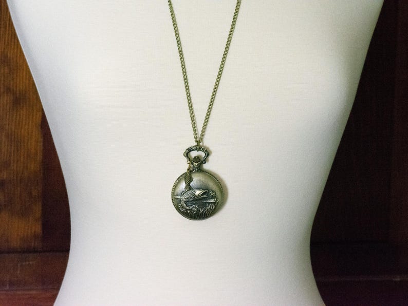 Pode incluir: Um colar dourado com um pingente de rel&oacute;gio de bolso redondo que apresenta um design de peixe detalhado.