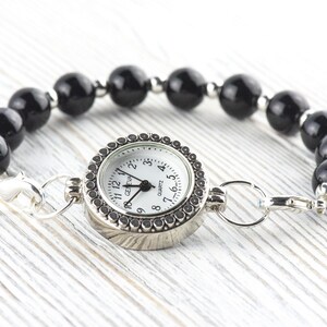 May include: A black beaded bracelet watch with a silver watch face. The watch face has a white background with black numerals and hands. The bracelet is made of large, round, black beads and silver metal accents.