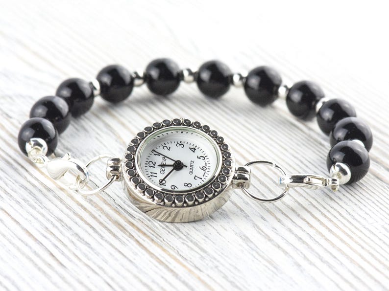 May include: A black beaded bracelet watch with a silver watch face. The watch face has a white background with black numerals and hands. The bracelet is made of large, round, black beads and silver metal accents.