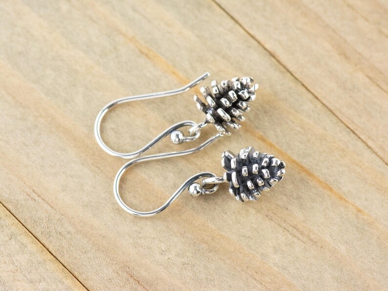 May include: A pair of silver earrings with a pine cone design. The earrings are dangle style with a small ball at the top of the hook.