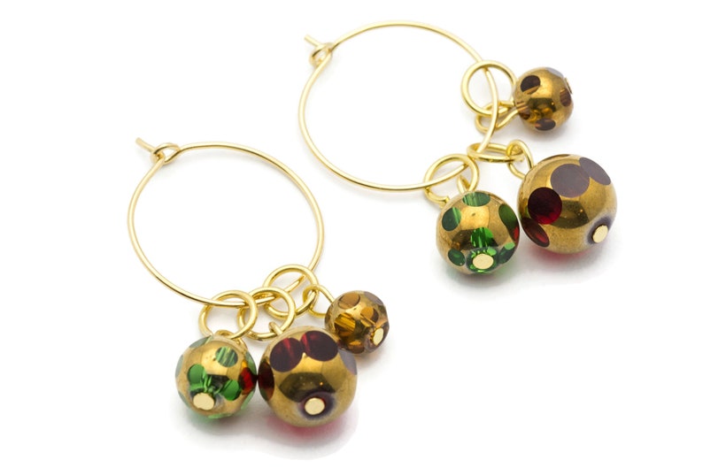 May include: Gold hoop earrings with three dangling charms. The charms are round and have a gold finish with green, red, and brown polka dots.