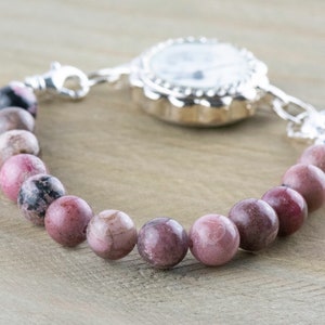 May include: A silver pocket watch with a pink and black beaded bracelet attached. The watch face is white with black markings.