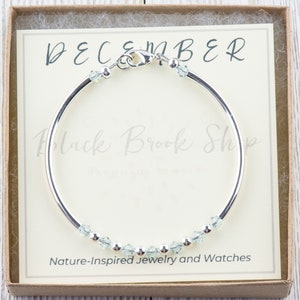 May include: A silver bangle bracelet with a clasp and light blue crystals. The bracelet is in a box with a white card that says "DECEMBER" and "Black Brook Shop - Nature-Inspired Jewelry and Watches".