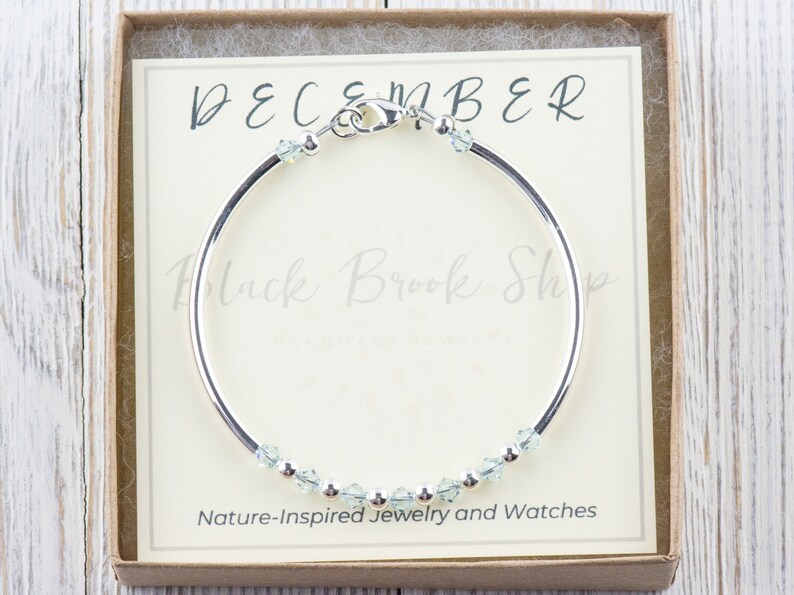 May include: A silver bangle bracelet with a clasp and light blue crystals. The bracelet is in a box with a white card that says "DECEMBER" and "Black Brook Shop - Nature-Inspired Jewelry and Watches".