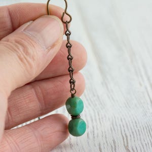 May include: A single earring with a bronze-colored chain and hook. Two turquoise-colored, cube-shaped beads are connected by a bronze-colored wire. The earring is held in a person's hand.