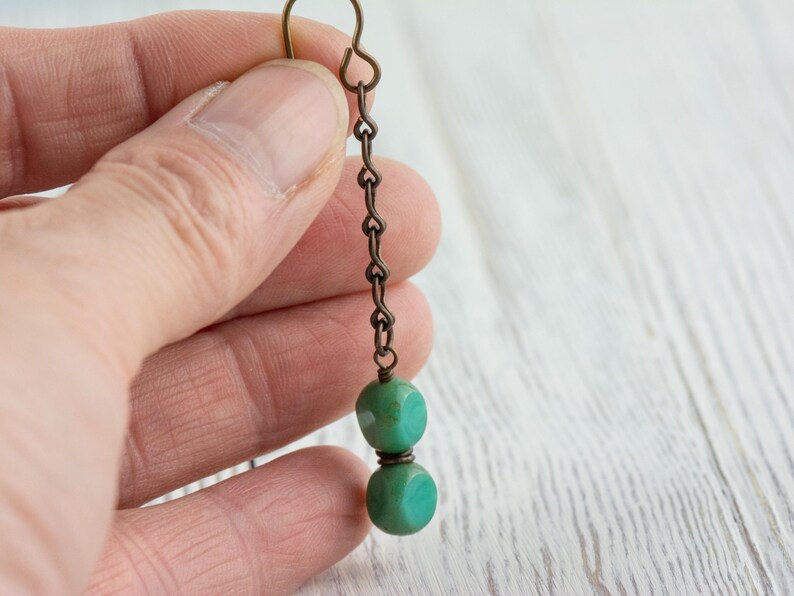 May include: A single earring with a bronze-colored chain and hook. Two turquoise-colored, cube-shaped beads are connected by a bronze-colored wire. The earring is held in a person's hand.