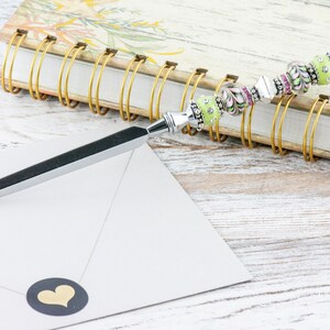 Peut inclure: Un ouvre-lettre en argent avec un manche en perles vertes et roses. Le manche est d&eacute;cor&eacute; de strass. L'ouvre-lettre repose sur un carnet ferm&eacute; avec une reliure spirale dor&eacute;e.